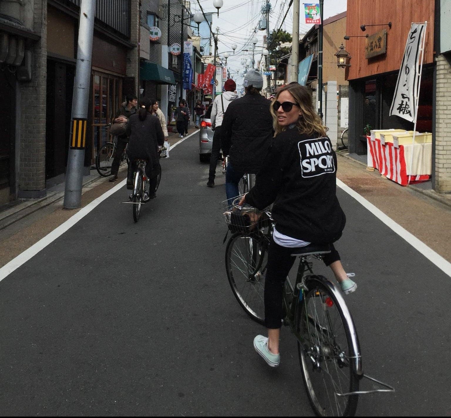 Ana Van Pelt riding a bicycle down a narrow city street, wearing a black hoodie with 'MILO SPORT' printed in white on the back, black pants, and light mint sneakers. Other cyclists and pedestrians are visible on the road between buildings with overhead wires.